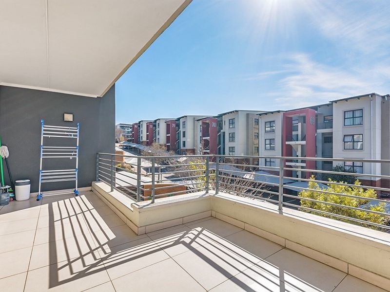 A spacious, tiled balcony featuring a metal balustrade and covered ceiling, offering an open view of the surrounding apartment complex.