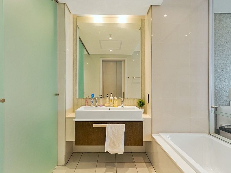 Modern bathroom featuring a floating vanity with a ceramic basin, large wall mirror, bathtub, and frosted glass door with neutral tiled finishes.