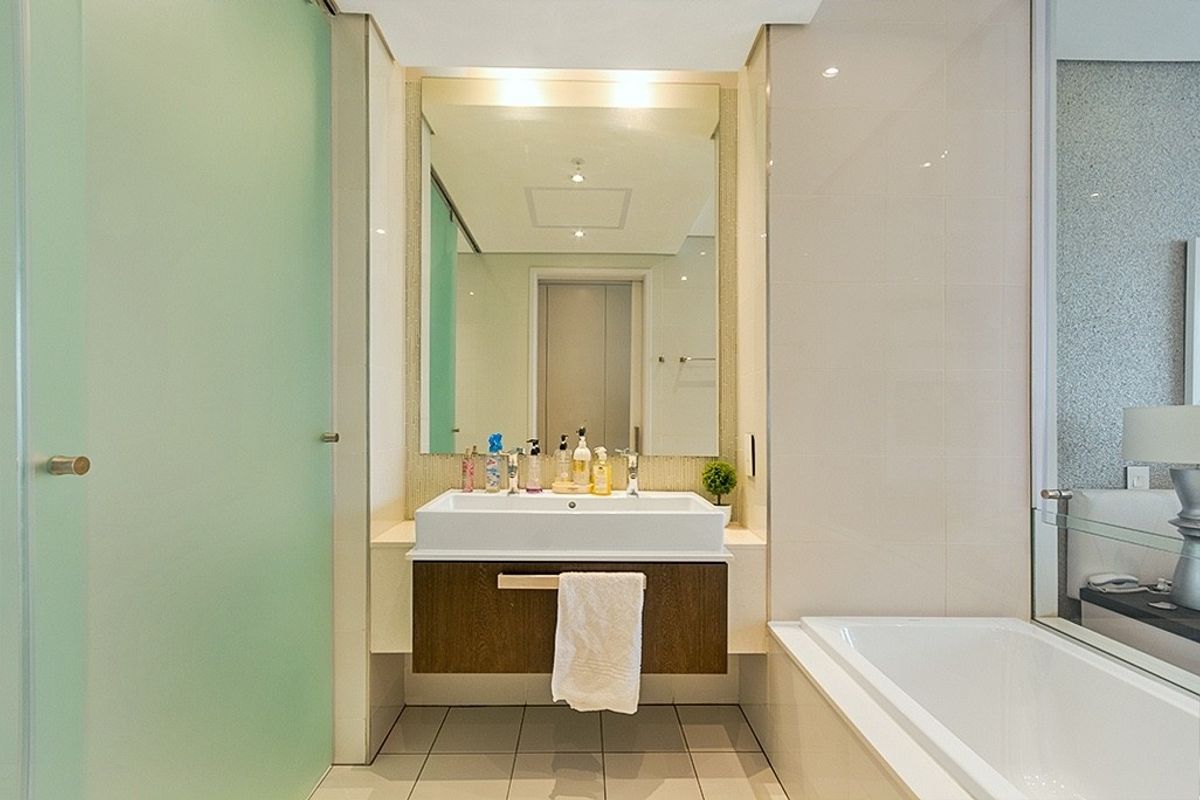 Modern bathroom featuring a floating vanity with a ceramic basin, large wall mirror, bathtub, and frosted glass door with neutral tiled finishes.