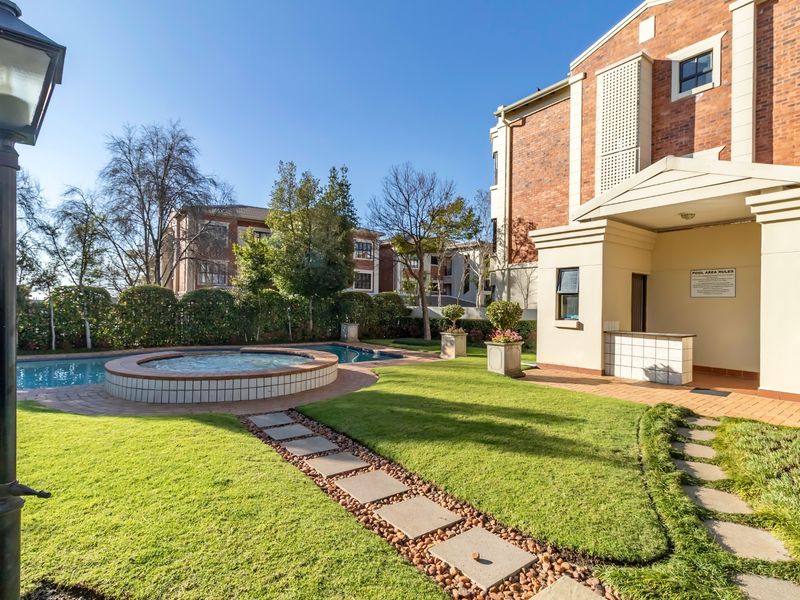 Well-maintained communal pool area featuring a raised spa, manicured lawn, paved pathways, and a covered entrance to the apartment building.