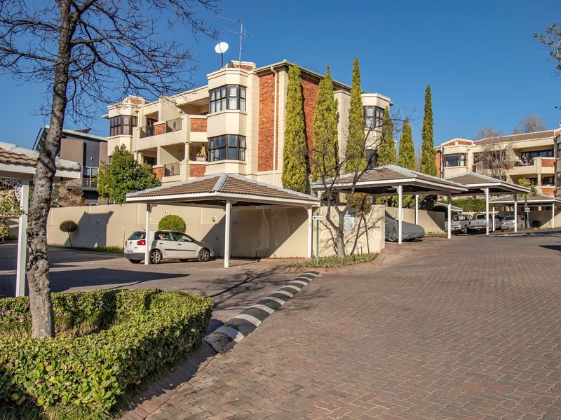 Exterior view of an apartment complex featuring covered parking carports, paved driveway, and multi-story brick and plaster building facade.