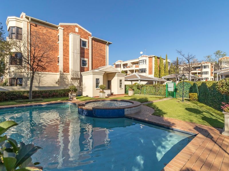 A well-maintained communal pool area featuring a raised spa, brick paving, and landscaped gardens set against a multi-story brick apartment building.