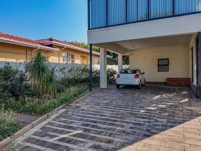 A modern residential exterior featuring a paved driveway, covered parking space, and a well-maintained landscaped garden with boundary wall and electric fencing.