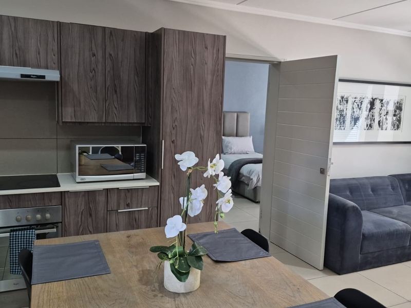 Modern open-plan kitchen featuring dark wood-grain cabinetry, integrated appliances, and tiled flooring, with seamless flow into the adjacent living area and bedroom.