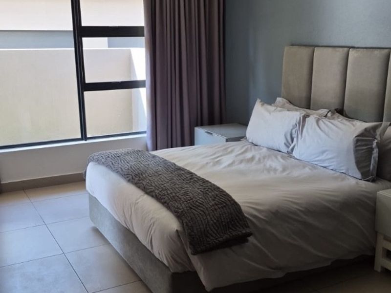 A well-lit bedroom featuring neutral tiled flooring, recessed ceiling lighting, and a large window with integrated curtain tracks.