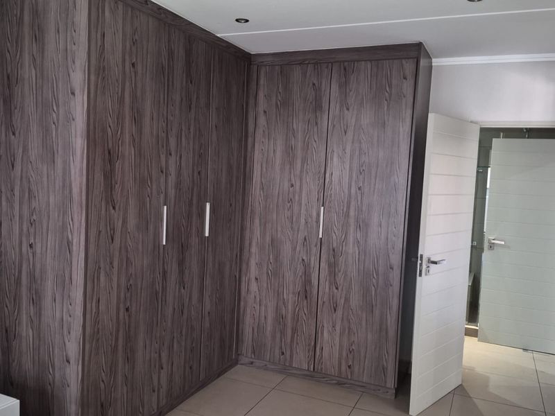 A bedroom featuring extensive dark wood-grain built-in cupboards, neutral tiled flooring, and recessed ceiling lighting, with an adjacent doorway leading to an en-suite.