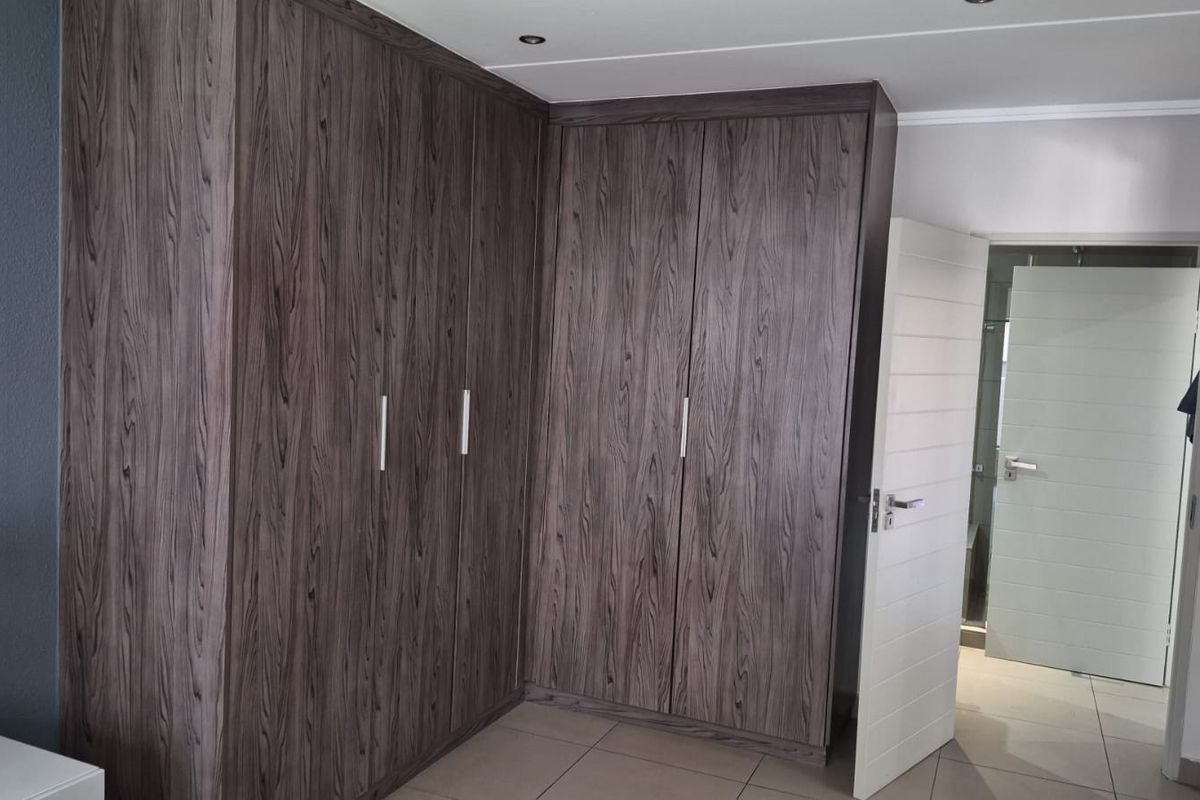 A bedroom featuring extensive dark wood-grain built-in cupboards, neutral tiled flooring, and recessed ceiling lighting, with an adjacent doorway leading to an en-suite.