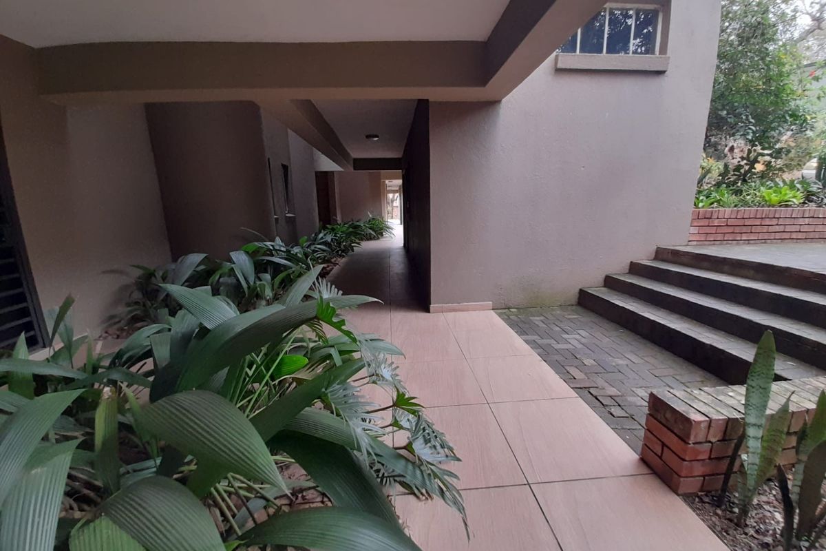 A covered outdoor walkway featuring tiled flooring, concrete steps, and brick retaining walls, leading towards a rear garden area.