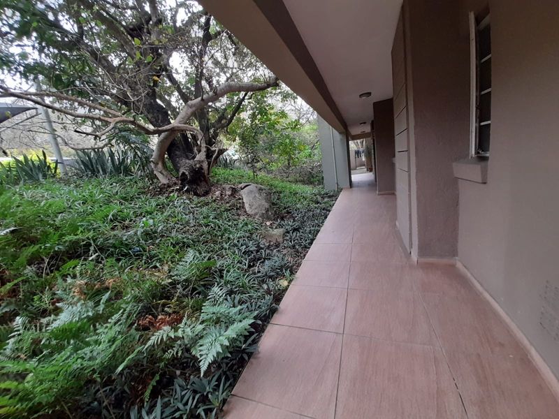 A covered outdoor patio or walkway featuring tiled flooring, adjacent to a landscaped garden area with mature trees.