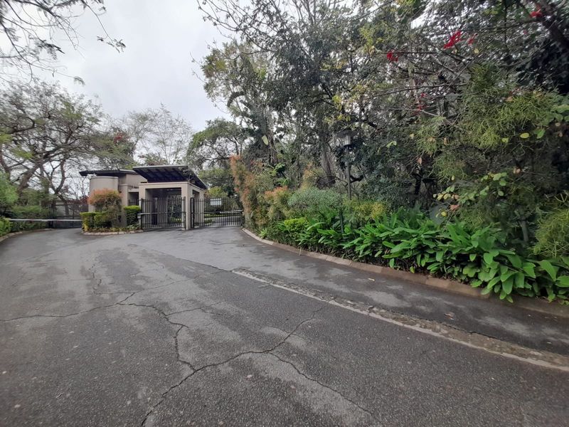 The entrance to the apartment complex features a secure gated access point with a guard house, surrounded by mature trees and landscaped greenery.