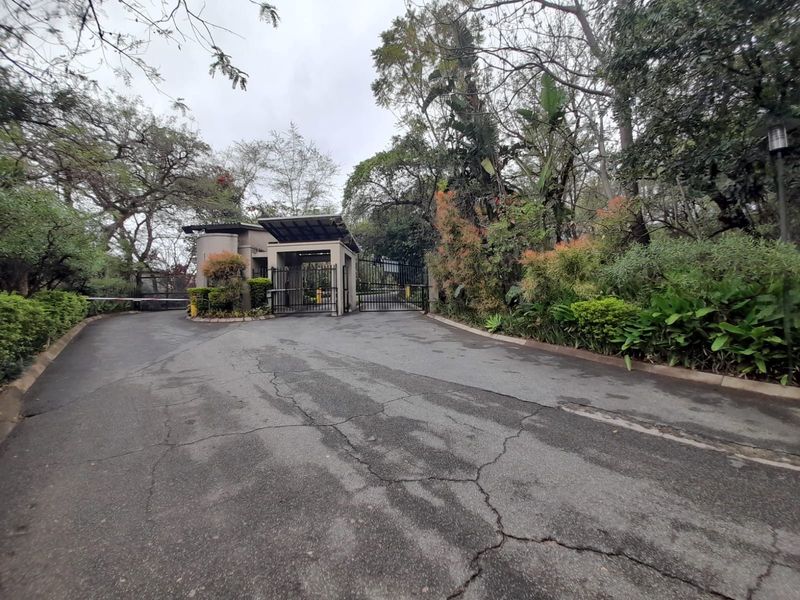 The entrance to the apartment complex features a security gate, boom barrier, and a guard house, surrounded by mature trees and landscaped greenery.