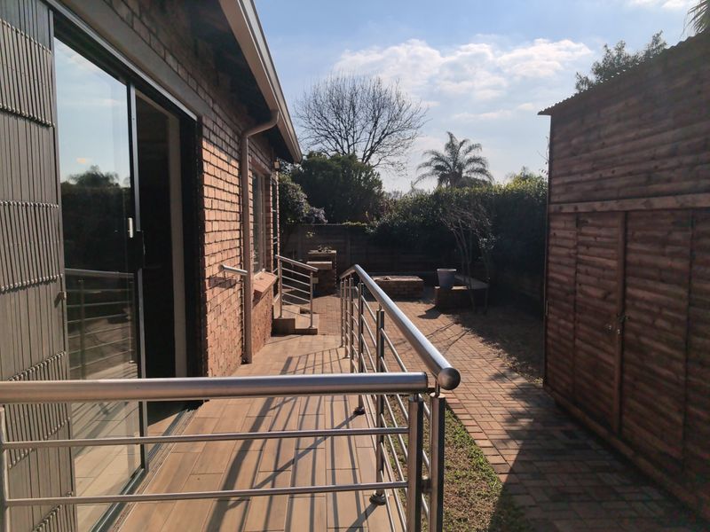 A well-maintained outdoor patio area featuring a stainless steel balustrade, tiled flooring, and a paved walkway leading to a built-in braai and storage shed.