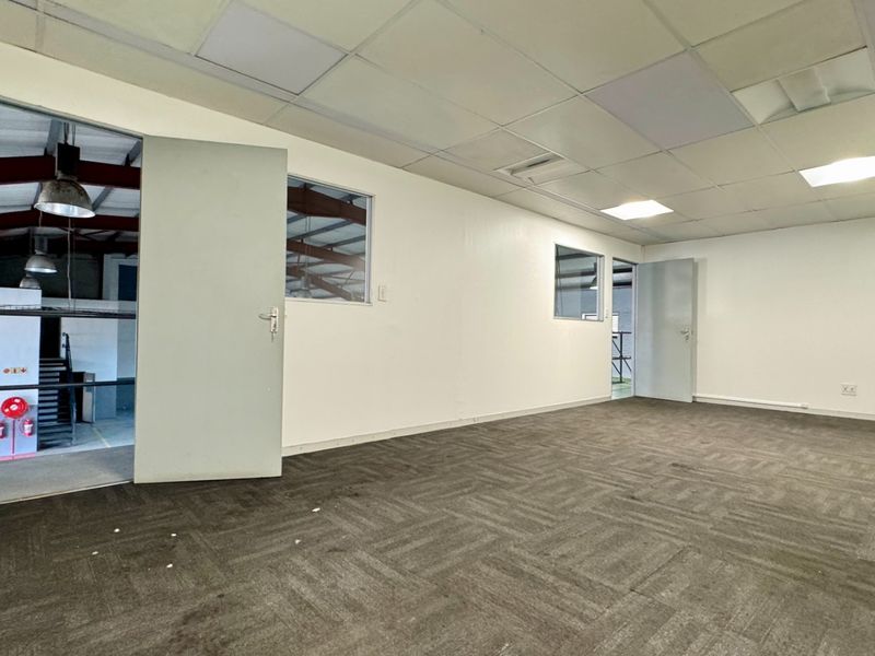 A vacant office space featuring carpet tiles, a suspended ceiling with recessed lighting, and a wall-mounted air conditioning unit, overlooking an industrial warehouse area.