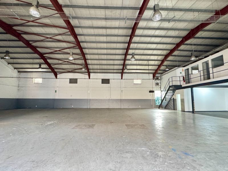 Spacious industrial warehouse featuring high ceilings with exposed steel trusses, concrete flooring, and a mezzanine level with metal staircase access.