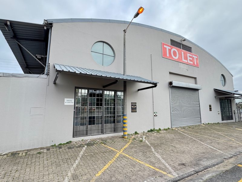 Front elevation of an industrial warehouse featuring a roller shutter door, metal-framed windows, and dedicated paved parking space.