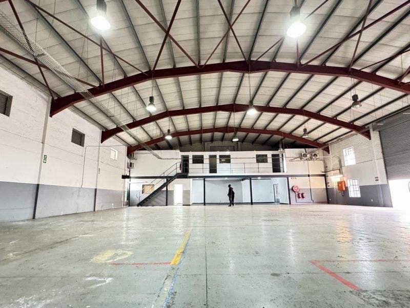 Spacious industrial warehouse featuring high ceilings with exposed steel trusses, a mezzanine level, concrete flooring, and a large roller shutter door for easy access.