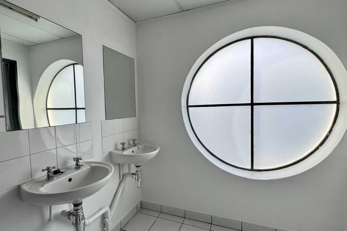 A functional industrial-style bathroom featuring dual wall-mounted washbasins, tiled splashbacks, and a distinctive circular window providing natural light.
