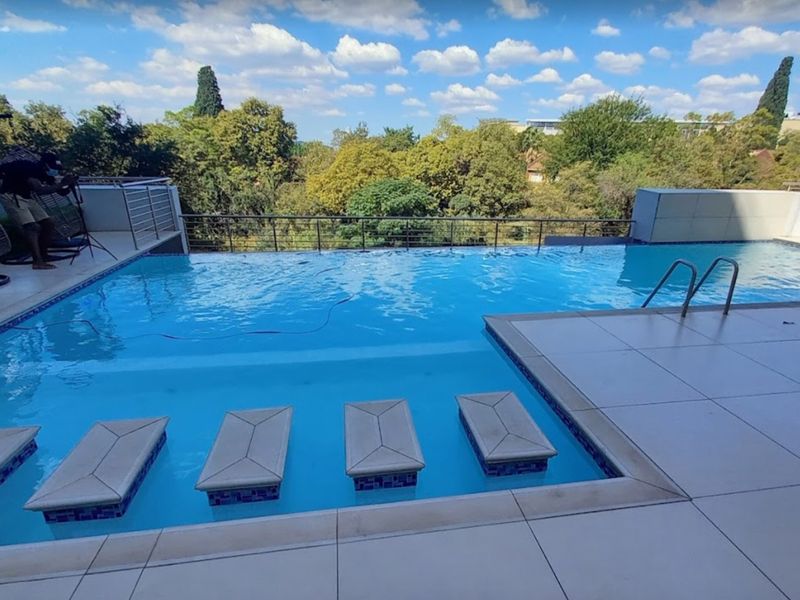 An elevated infinity edge swimming pool featuring built-in in-pool seating, tiled decking, and a metal balustrade overlooking a lush green landscape.