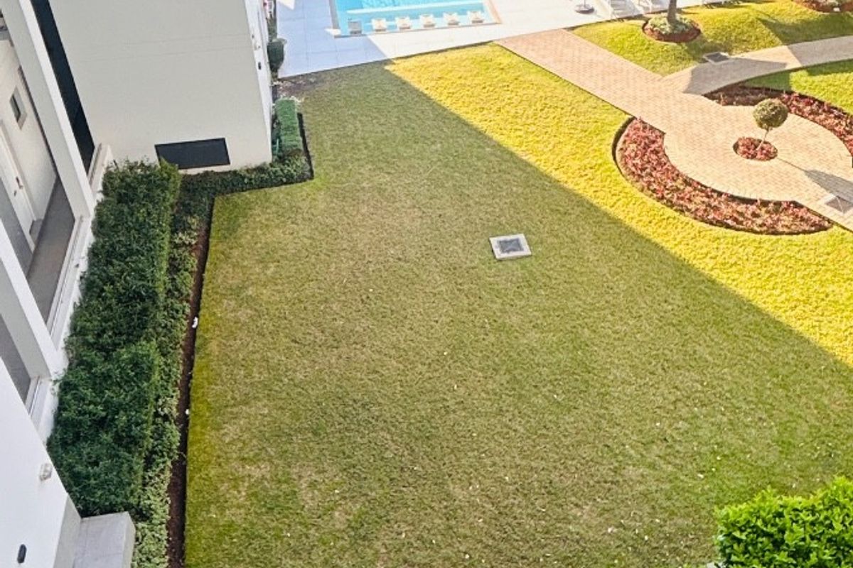 An elevated aerial view of the apartment complex grounds, featuring a well-maintained swimming pool, landscaped lawn, and paved garden pathways.