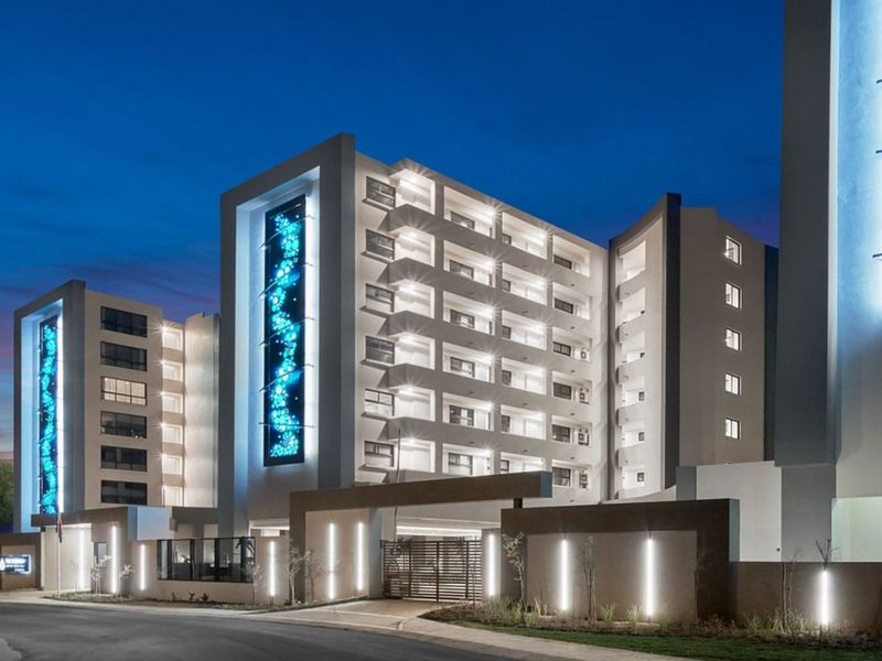 A modern multi-story apartment building exterior at dusk, featuring contemporary architectural design, illuminated decorative panels, and a secure gated entrance.