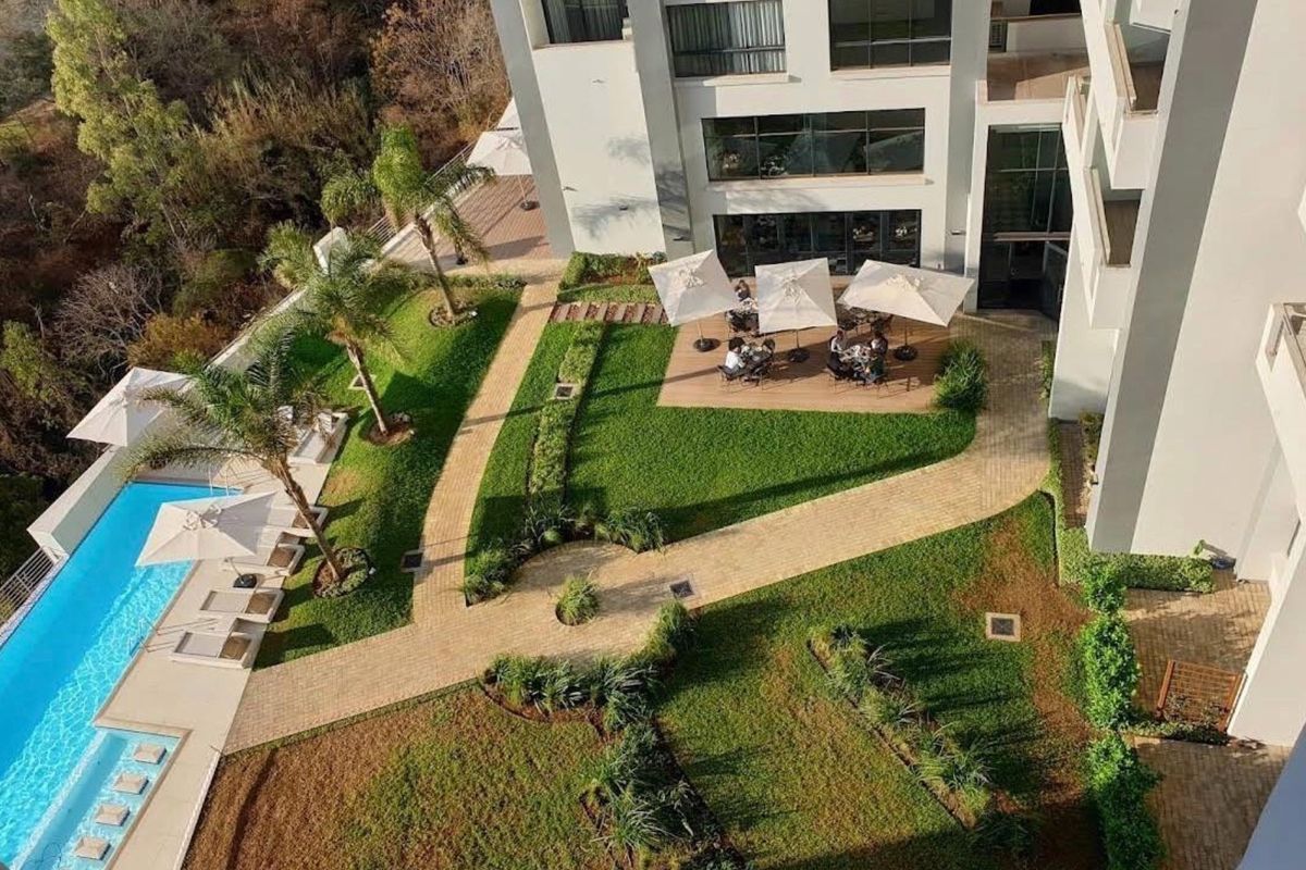 An elevated aerial view of the apartment complex grounds, featuring a swimming pool, landscaped gardens, paved pathways, and an outdoor seating area.