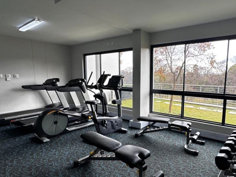 A well-lit apartment gym featuring large windows with garden views, carpeted flooring, and a selection of exercise equipment.
