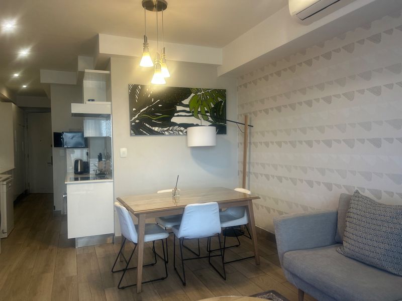 Open-plan living and dining area featuring wood-look tile flooring, modern pendant lighting, and a compact integrated kitchen space with an air conditioning unit.