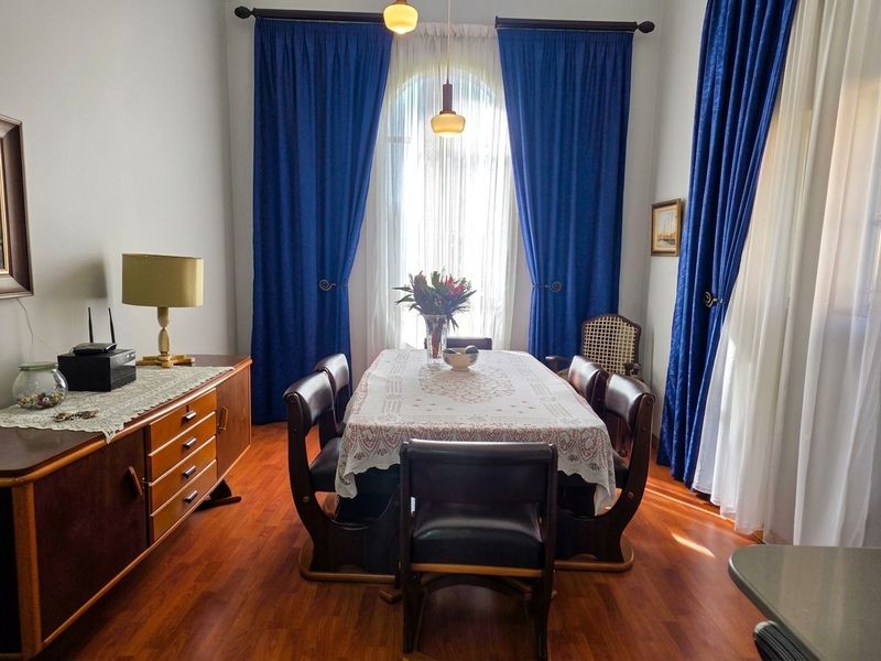 A formal dining room featuring warm laminate flooring, an elegant arched window with floor-to-ceiling curtains, and classic pendant lighting.