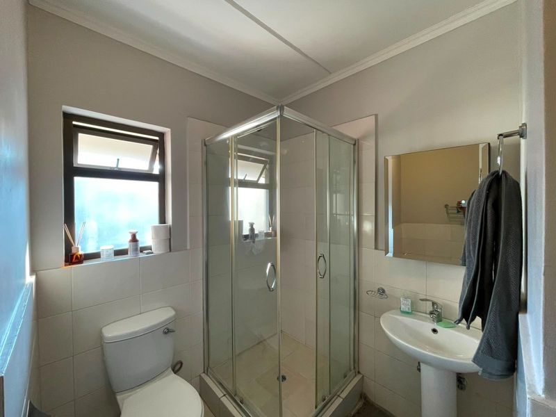 A functional bathroom featuring a glass shower enclosure, pedestal basin, toilet, and wall-mounted mirror with natural light from a side window.