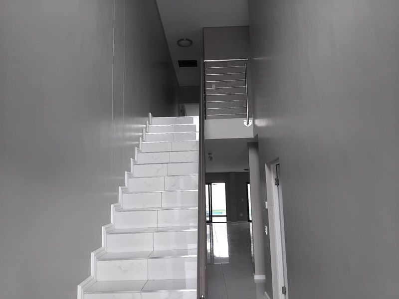 A bright, modern hallway featuring a tiled staircase with a stainless steel balustrade and polished floor tiles leading to an open living area.