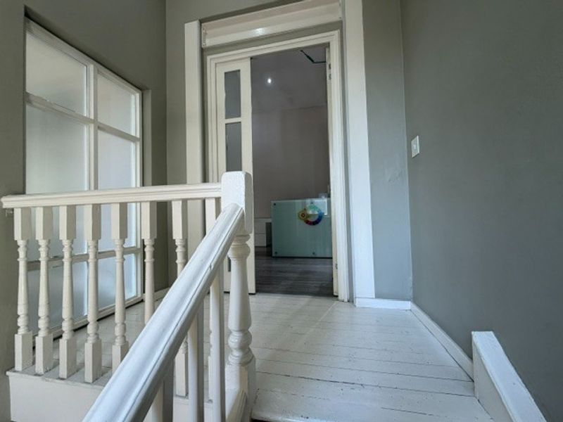 A bright hallway featuring a white wooden balustrade, painted timber flooring, and neutral grey walls leading into an adjacent office space.