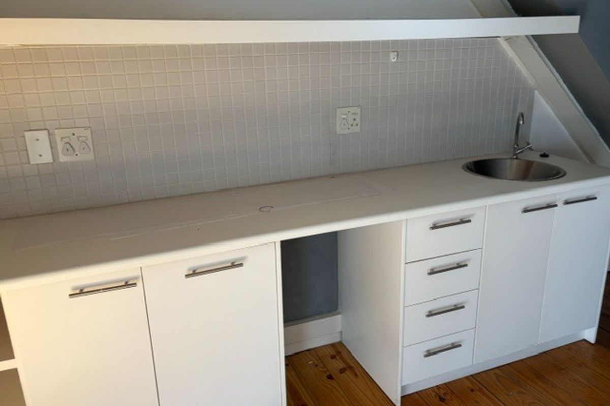 A clean, compact kitchenette featuring white built-in cabinetry, a stainless steel sink, tiled backsplash, and a floating shelf under a sloped ceiling.