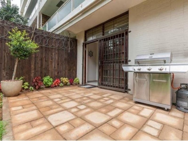A private paved patio area featuring a secure entrance gate, timber boundary fencing with security wire, and direct access to the apartment interior.