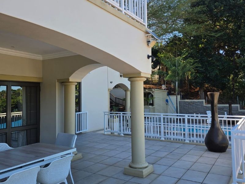 Spacious covered patio featuring tiled flooring, arched columns, and a white balustrade railing, overlooking the pool area and garden.