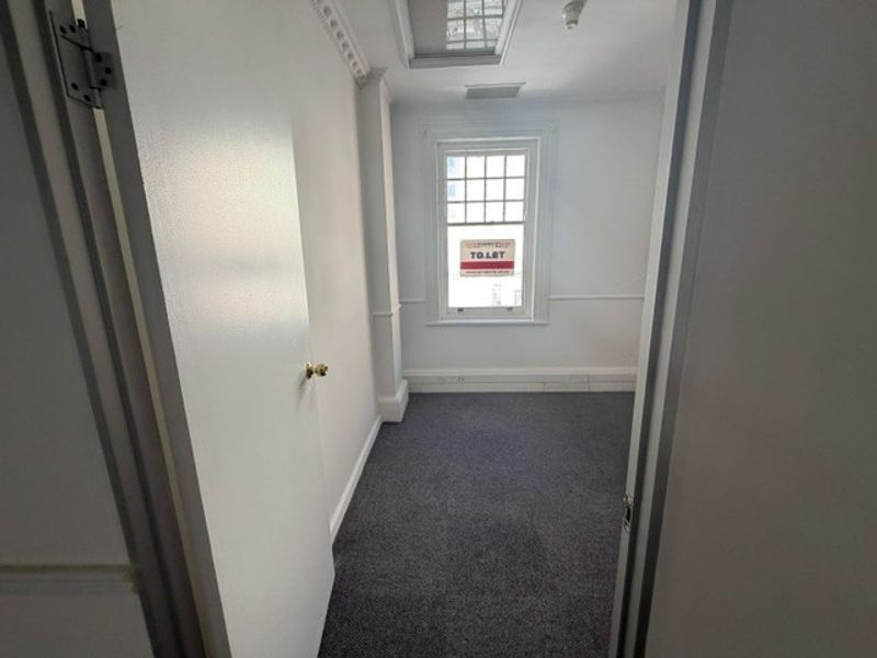 A bright, empty office space featuring neutral walls, carpeted flooring, classic chair rail molding, and a large sash window providing natural light.