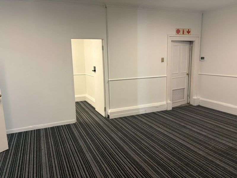 A clean, empty office space featuring striped carpet flooring, chair rail molding, and a secondary access door with integrated ventilation.