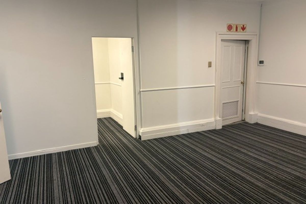 A clean, empty office space featuring striped carpet flooring, chair rail molding, and a secondary access door with integrated ventilation.