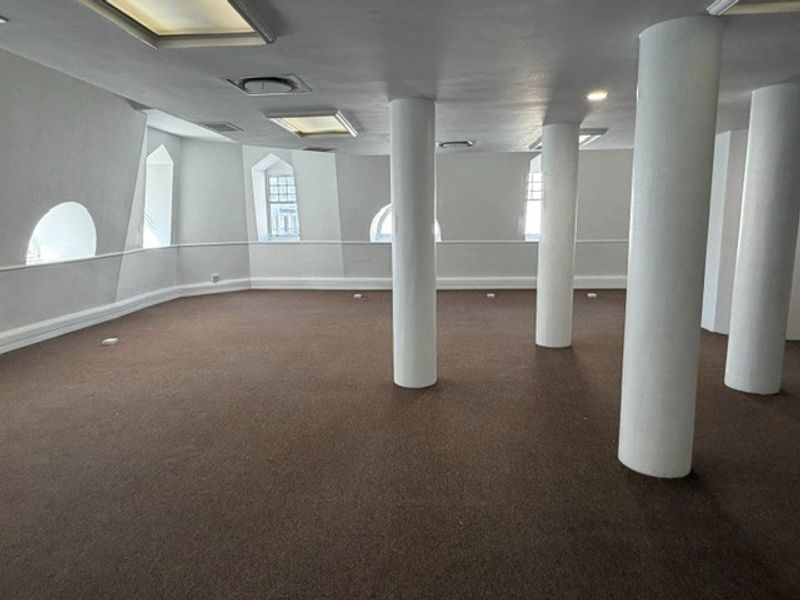 An open-plan office space featuring structural columns, carpeted flooring, and multiple arched windows providing natural light.