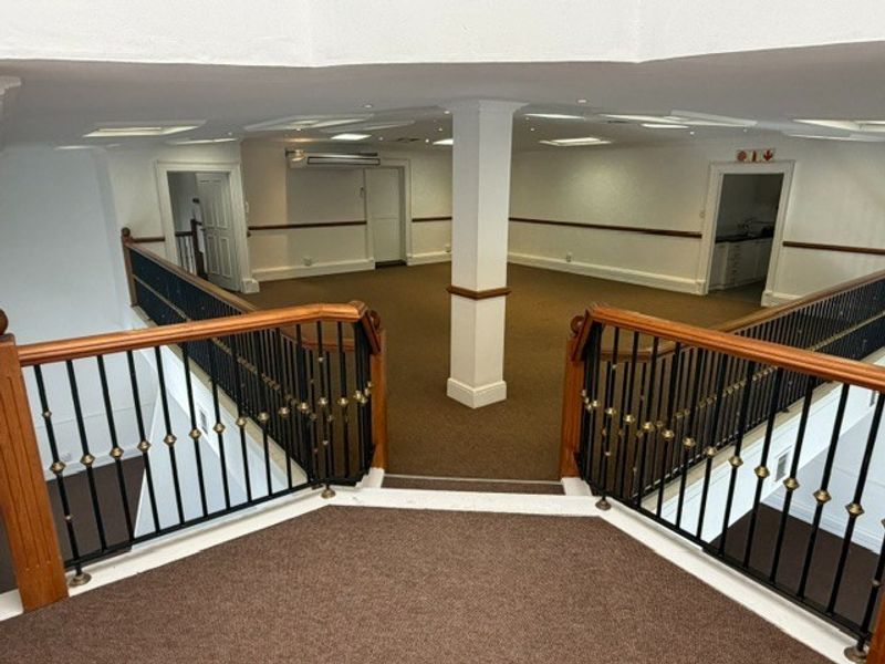 Spacious open-plan office area featuring a central support column, carpeted flooring, and a decorative wrought iron balustrade overlooking the lower level.