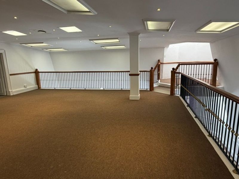 Spacious open-plan office mezzanine area featuring carpeted flooring, recessed ceiling lighting, and a secure metal balustrade with wooden handrails.