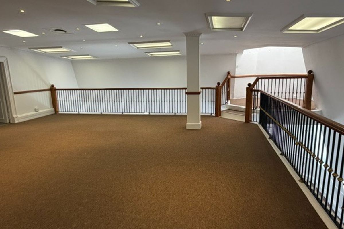 Spacious open-plan office mezzanine area featuring carpeted flooring, recessed ceiling lighting, and a secure metal balustrade with wooden handrails.