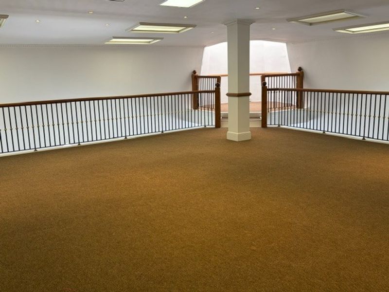 An open-plan office mezzanine area featuring carpeted flooring, recessed ceiling lights, and a decorative metal balustrade with a wooden handrail.