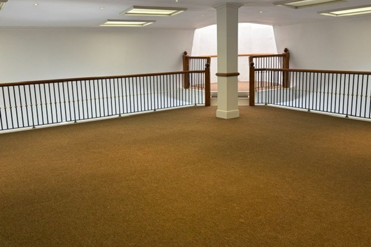 An open-plan office mezzanine area featuring carpeted flooring, recessed ceiling lights, and a decorative metal balustrade with a wooden handrail.