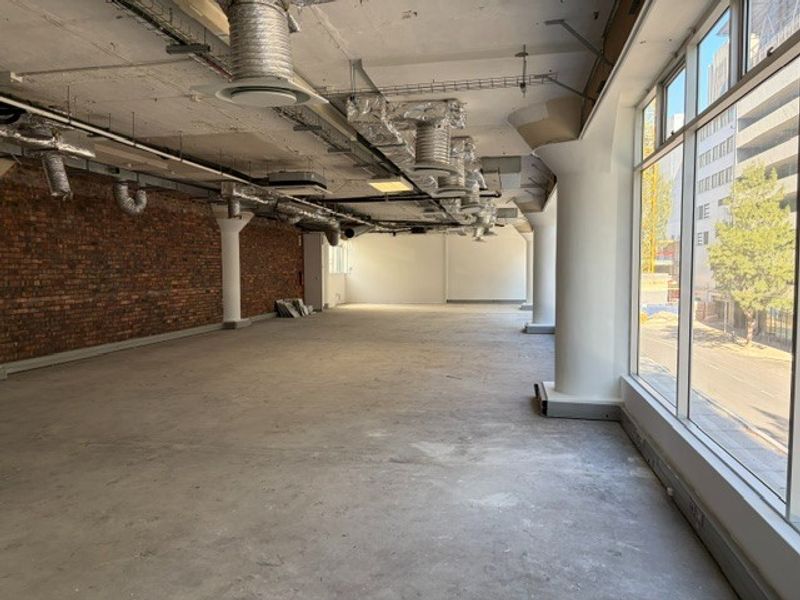 An expansive, open-plan office space featuring industrial-style exposed brick walls, concrete floors, and large windows providing ample natural light.