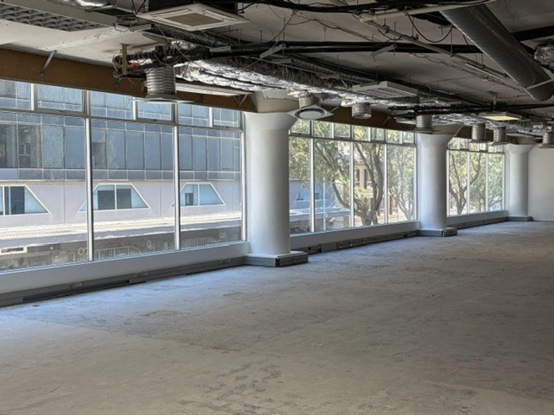 An expansive, open-plan office space featuring large windows, exposed industrial ceiling infrastructure, and structural concrete columns, currently in a shell condition.