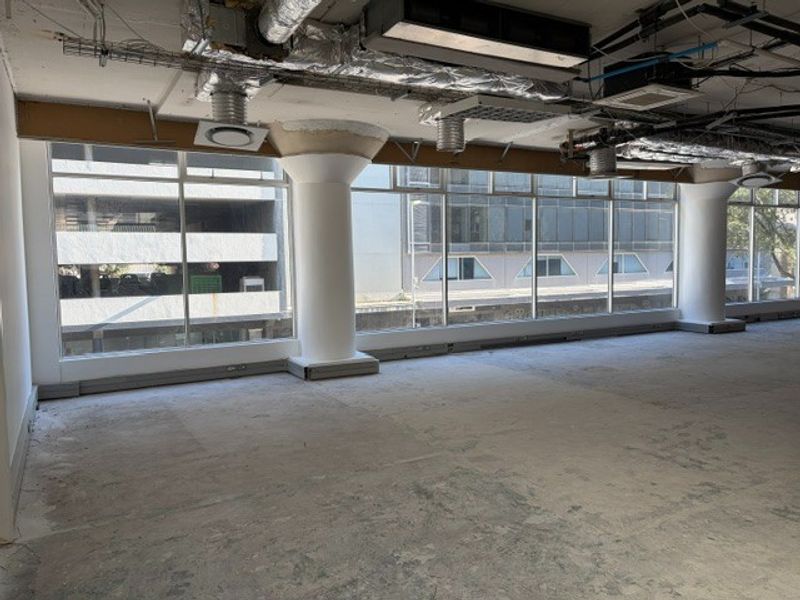 An open-plan office space featuring large windows, exposed industrial ceiling infrastructure, and concrete flooring, currently in a shell condition ready for fit-out.
