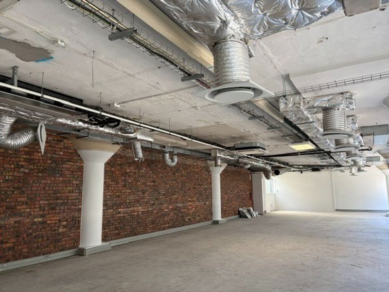 An open-plan office space featuring industrial-style exposed brick walls, concrete columns, and exposed ceiling infrastructure with HVAC ducting.