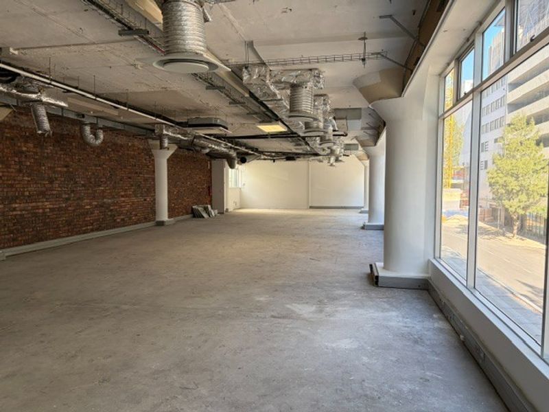Spacious open-plan office area featuring industrial-style exposed brick walls, exposed ceiling ducting, and large windows providing ample natural light.