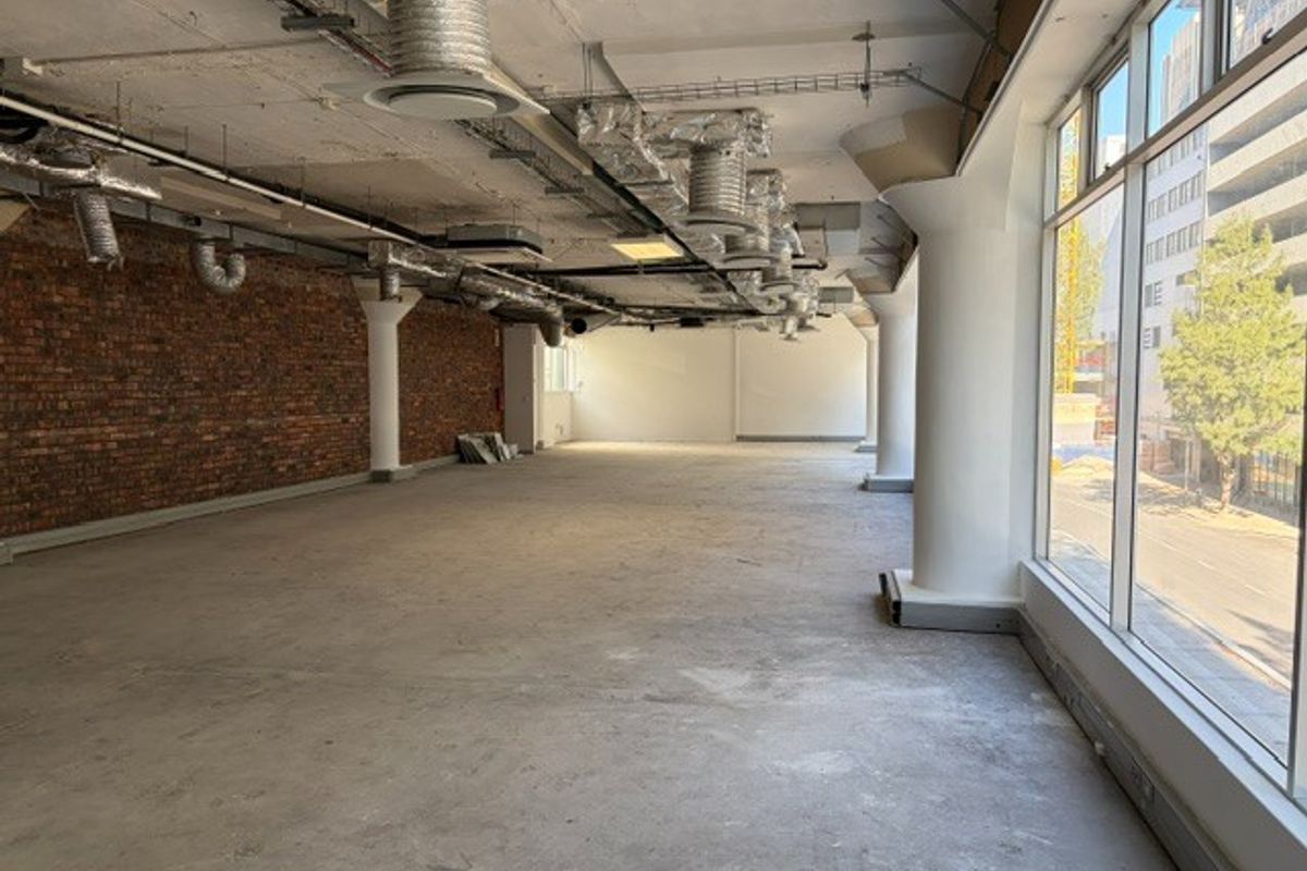 Spacious open-plan office area featuring industrial-style exposed brick walls, exposed ceiling ducting, and large windows providing ample natural light.