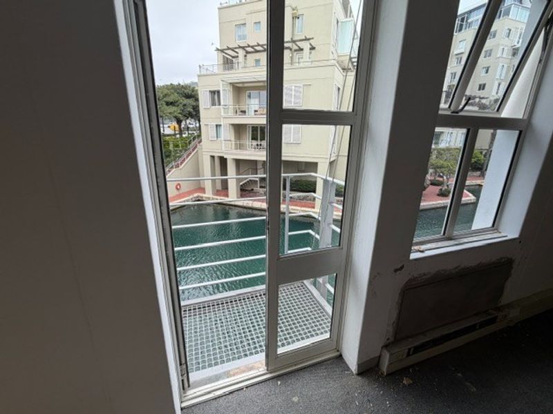 A small balcony with metal railing overlooking a canal, accessible via a sliding glass door in an office space with worn finishes.